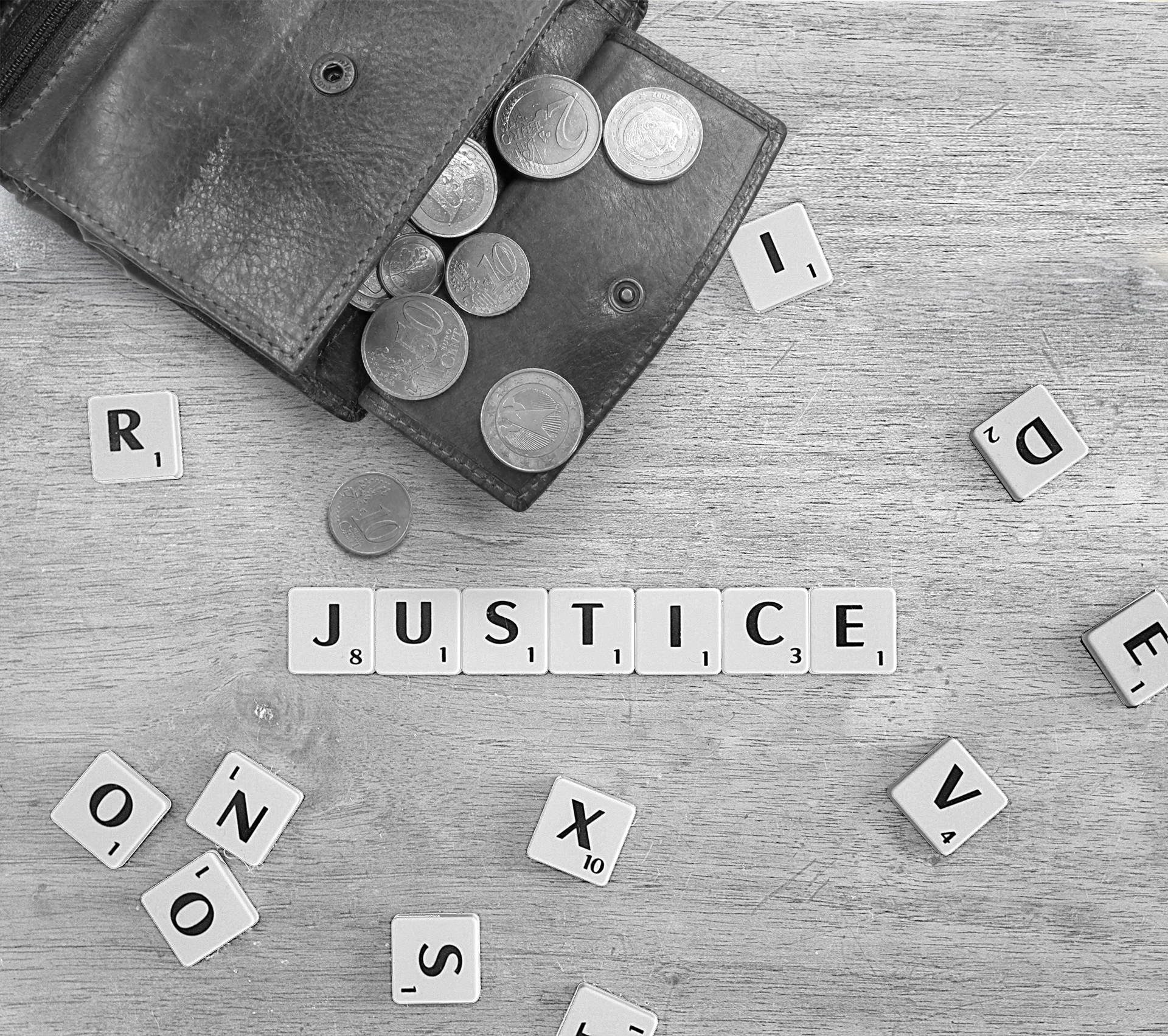 Schwarz-Weiß Aufnahme in der Draufsicht einer Geldbörse  mit offenem Kleingeldfach auf einem Holztisch mit einigen Scrabble Steinen, aus denen das Wort JUSTICE, also Gerechtigkeit zusammengelegt ist.